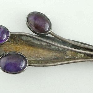 May include: A silver-toned brooch with three oval, deep purple amethyst stones. The brooch has a leaf-like design with a central, elongated element and two stems. The stones are set in a bezel setting.