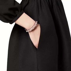 Puede incluir: Un vestido negro con un corpi&ntilde;o ajustado y una falda completa. El vestido tiene un escote en V y mangas largas. Una mujer lleva el vestido y tiene una pulsera de tachuelas plateadas y rosadas en la mu&ntilde;eca.