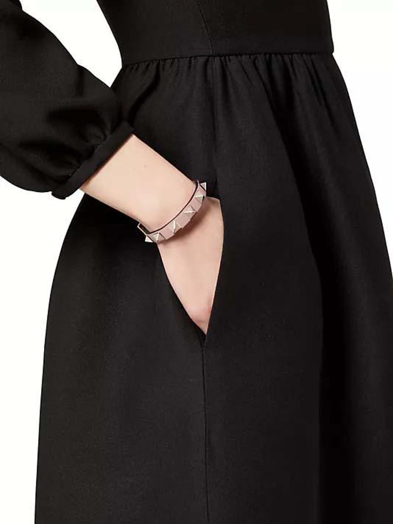 Puede incluir: Un vestido negro con un corpi&ntilde;o ajustado y una falda completa. El vestido tiene un escote en V y mangas largas. Una mujer lleva el vestido y tiene una pulsera de tachuelas plateadas y rosadas en la mu&ntilde;eca.