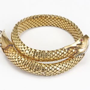 Könnte beinhalten: Ein goldfarbenes Schlangenarmband mit detailliertem Schuppenmuster. Der Schlangenkopf hat ein rotes Edelsteinauge und einen Verschluss. Das Armband ist gewunden und zeigt sein flexibles Design und seine aufwendige Verarbeitung. Schmuck.