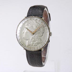 May include: A silver-toned wristwatch featuring a Morgan silver dollar coin as the watch face. The coin displays a profile of a woman and the year 1878. The watch has gold-colored hands and a black leather band.
