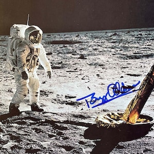 Op de afbeelding: Een zwart-witfoto van een astronaut op de maan, gesigneerd door Buzz Aldrin. De astronaut draagt een wit ruimtepak en staat naast een maanlander.