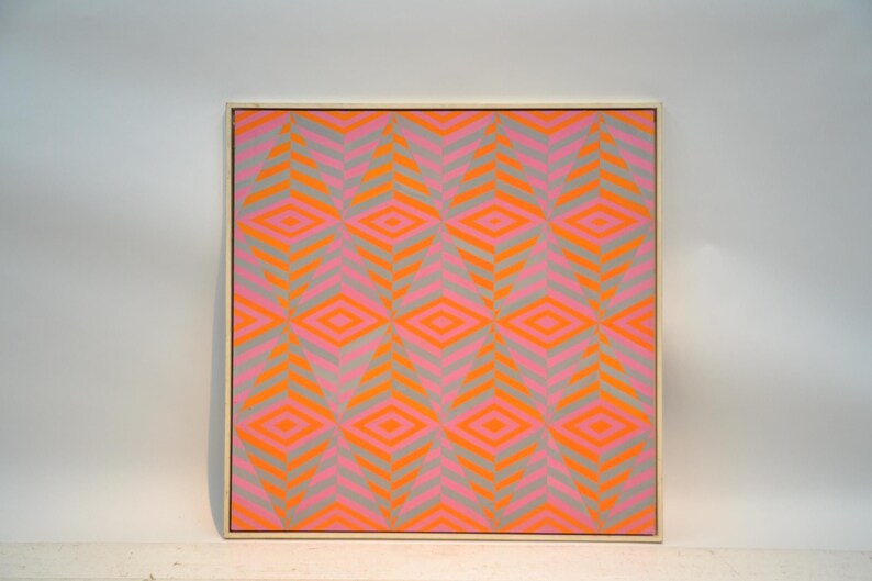 May include: Framed artwork with a geometric pattern. The design features repeating diamond shapes in vibrant orange and pink, set against a grey background. The artwork is encased in a light-coloured wooden frame.