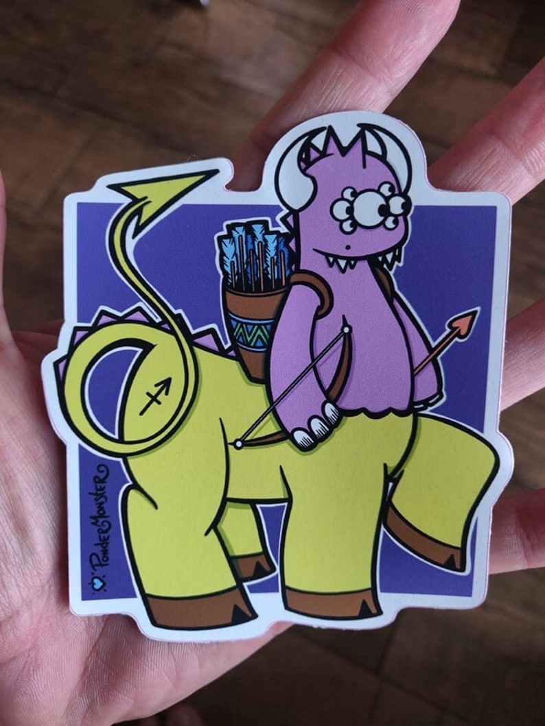 May include: A purple monster with three eyes riding a yellow centaur with a bow and arrow. The background is purple. The sticker says "♡ Powder Monster"
