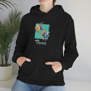 Könnte beinhalten: Schwarzer Kapuzenpullover mit einem türkisfarbenen Quadrat mit zwei Cartoon-Fischen und dem Wort "Pisces". Die Fische sind orange und blau mit skurrilen Details. Der Pullover hat eine Vordertasche und eine Kapuze mit Kordelzug.