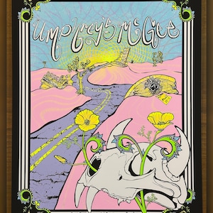 Könnte beinhalten: Ein farbenfrohes Konzertplakat für Umphrey's McGee, das eine Wüstenlandschaft mit einem Schädel und gelben Blumen zeigt. Das Plakat hat einen schwarzen Rahmen mit dekorativen Elementen und Text.