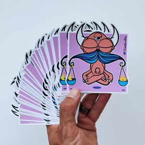 Peut inclure: Une pile d'autocollants roses et blancs repr&eacute;sentant une illustration de dessin anim&eacute; d'une cr&eacute;ature rose avec une moustache bleue et des cornes, assise en position de yoga sur une balance avec une lune croissante et un soleil de chaque c&ocirc;t&eacute;. L'autocollant porte le texte "PinkCloudFire" dans le coin sup&eacute;rieur droit.