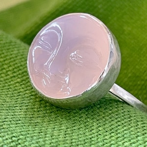 May include: A silver ring with a pink moonstone cabochon set in a bezel setting. The moonstone has a carved face design.