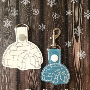May include: Two leather keychains shaped like igloos. The larger keychain is white with a silver outline and the smaller keychain is blue with a silver outline. Both keychains have a silver metal ring.