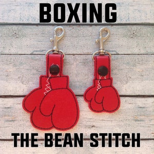 Könnte beinhalten: Zwei rote Boxhandschuh-Schlüsselanhänger mit silbernen Schlüsselringen. Die Schlüsselanhänger sind mit weißen Nähten bestickt und haben schwarze Knöpfe. Der Text "BOXING" befindet sich oben im Bild und "THE BEAN STITCH" unten.