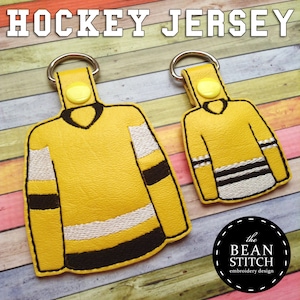 Può includere: Due portachiavi a forma di maglia da hockey gialla con strisce nere e bianche. Il portachiavi più grande ha una striscia bianca in basso e il portachiavi più piccolo ha tre strisce nere e bianche in basso. I portachiavi sono su uno sfondo di legno.