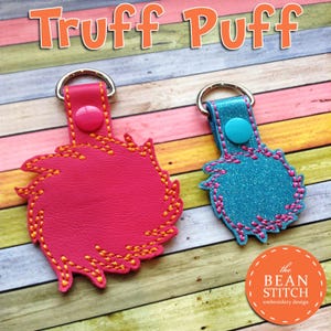 May include: Two Truff Puff keychains, one pink and one blue, with a yellow stitched design. The keychains are on a wooden background with the text "the BEAN STITCH embroidery design".