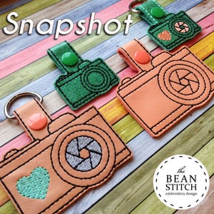 May include: Three embroidered camera keychains with a heart and lens design. The keychains are in green, orange, and peach colors. The text "the BEAN STITCH embroidery design" is visible in the bottom right corner.