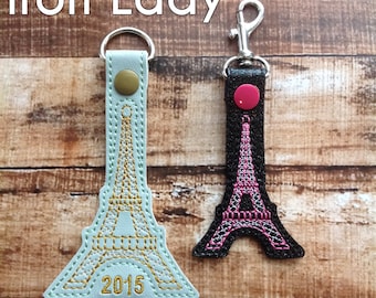 May include: Two Eiffel Tower keychains, one with a light blue background and gold embroidery, the other with a black background and pink embroidery. Both keychains have a silver metal ring and a button on the top. The light blue keychain has the year 2015 embroidered on the bottom.