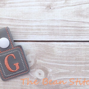 Op de afbeelding: Een grijze leren sleutelhanger met een witte knop en een vierkant patch met een gestikte koperkleurige letter G.