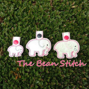 Può includere: Tre ornamenti a forma di elefante in feltro bianco con bottoni rosa. Gli elefanti sono di dimensioni diverse e presentano dettagli ricamati. Il testo "The Bean Stitch" è visibile sotto gli elefanti.