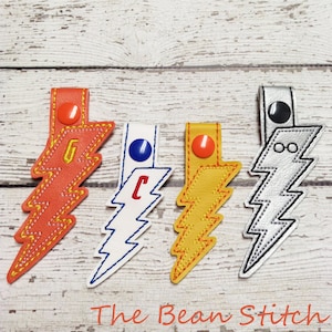 May include: Four lightning bolt shaped keychains with different colored fabric and embroidered letters. The keychains are orange, white, yellow, and silver.