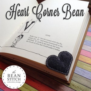 以下が含まれることがあります： グレーのフェルト製のハート型のしおりが付いた本のクローズアップ。本は「Heart Corner Bean」というタイトルと、愛についての短い詩が書かれたページを開いています。本は木製の表面の上に置かれています。