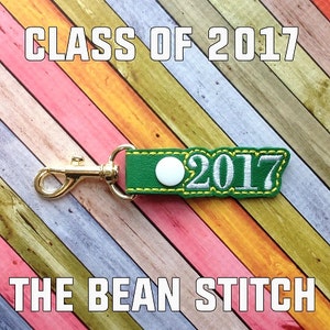 May include: Green leather keychain with a white button and the text "2017" in white embroidery. The keychain has a gold metal clasp. The text "CLASS OF 2017" and "THE BEAN STITCH" are visible in the background.