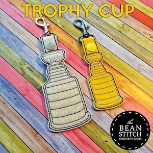 Könnte beinhalten: Zwei gestickte Trophäen-Schlüsselanhänger, einer weiß und einer gelb, mit silbernen Schlüsselringen. Die Schlüsselanhänger befinden sich auf einem Holzgrund mit bunten Streifen.