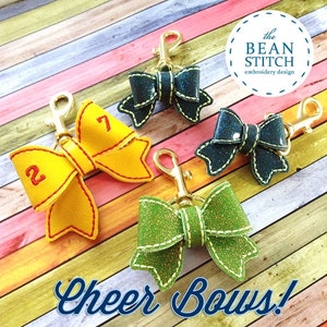 Può includere: Tre fiocchi da cheerleader con portachiavi dorati. I fiocchi sono gialli, verdi e blu con glitter. I fiocchi sono decorati con i numeri 2 e 7. Il testo "the BEAN STITCH embroidery design" è nell'angolo in alto a destra dell'immagine. Il testo "Cheer Bows!" è nella parte inferiore dell'immagine.