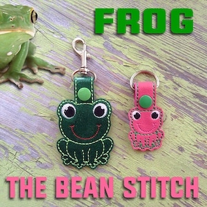 Könnte beinhalten: Zwei fröschchenförmige Schlüsselanhänger, einer grün und einer rosa, mit gestickten Details und einem lächelnden Gesicht. Die Schlüsselanhänger sind an Metallringen befestigt und haben einen Knopf auf der Rückseite. Das Wort "FROG" ist in grünen Buchstaben über den Schlüsselanhängern geschrieben. Der Text "THE BEAN STITCH" ist in rosa Buchstaben unter den Schlüsselanhängern geschrieben.