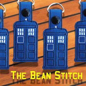 May include: Three blue and white Tardis keyrings with a silver ring. The keyrings are on a wooden surface. The text "THE BEAN STITCH" is at the bottom of the image.