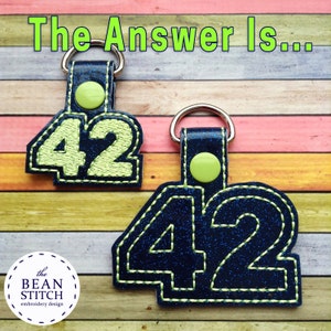 May include: Two blue and green keychains with the number 42 embroidered on them. The keychains are on a wooden background with the text "The Answer Is..." above them.
