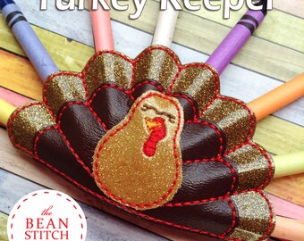 Puede incluir: Un porta lápices con forma de pavo marrón y dorado con un pico y ojos rojos. El porta lápices está decorado con purpurina y tiene un respaldo de fieltro marrón. El texto "Turkey Keeper" está escrito en letras blancas sobre el porta lápices.