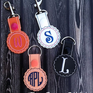 May include: Four personalized keychains with different colored leather straps and embroidered initials. The keychains feature a round, scalloped design with a single initial in the center. The initials are W, S, L, and APL. The keychains are displayed on a wooden background.