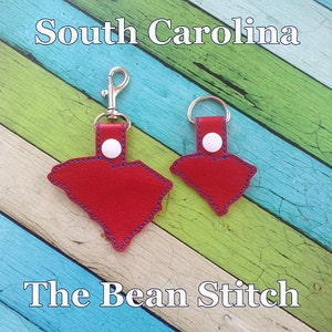 Puede incluir: Dos llaveros de cuero rojo con forma del estado de Carolina del Sur. Los llaveros tienen un botón blanco y un anillo de metal plateado. El llavero más grande tiene un cierre de metal plateado. Los llaveros están sobre un fondo de madera con el texto "South Carolina" y "The Bean Stitch".