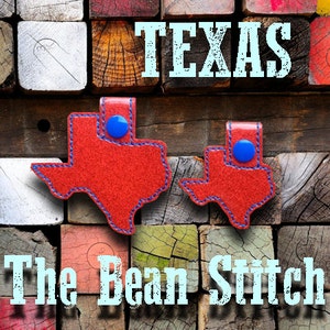 May include: Two red leather Texas-shaped keychains with blue stitching and blue button accents. The keychains are displayed on a wooden background with the text "TEXAS" and "The Beam Stitch" in blue.