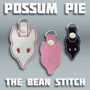 May include: Three embroidered keychains shaped like fox heads. The keychains are white, pink, and black. The text "Possum Pie" and "The Bean Stitch" are visible at the top and bottom of the image.