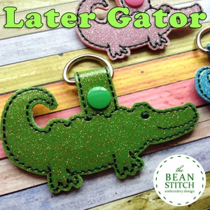 May include: Green glitter alligator keyring with a green button and the text "Later Gator" in yellow letters. The keyring is on a wooden background.