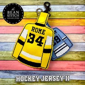 Peut inclure: Porte-clés représentant un maillot de hockey jaune avec le nom "ROME" et le numéro "34" en noir. Un porte-clés bleu en forme de maillot de hockey est visible en arrière-plan. Le texte "HOCKEY JERSEY II" est en bas.