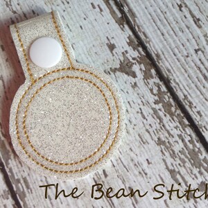 May include: A white glitter circle with a gold stitched border, attached to a white strap with a white button. The circle is blank and ready for personalization. The Bean Stitch is printed below the circle.