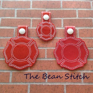 Può includere: Tre portachiavi in feltro rosso a forma di casco da pompiere con cuciture bianche e un bottone bianco. I portachiavi sono disposti in fila dal più grande al più piccolo. Il testo "The Bean Stitch" è visibile nella parte inferiore dell'immagine.