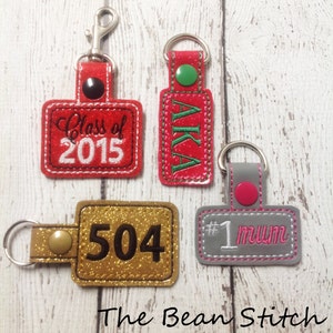 May include: Four different keychains with various designs. The first keychain is red with glitter and says "Class of 2015". The second keychain is red with glitter and says "AKA". The third keychain is gold with glitter and says "504". The fourth keychain is grey with glitter and says "#1 mum".