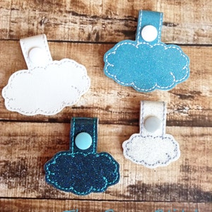 May include: Four cloud-shaped fabric keychains with different colors and textures. Two are white, one is teal blue with glitter, and one is dark blue with glitter. Each keychain has a loop at the top with a button.