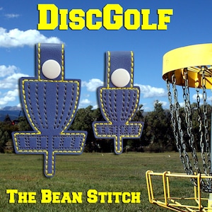 May include: Two blue leather disc golf keychain charms with yellow stitching, featuring a disc golf basket design. The charms are displayed against a backdrop of a blue sky and a disc golf basket.