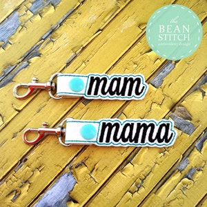 May include: Two white keychains with the words "mam" and "mama" in black script. Each keychain has a teal accent and a silver clasp. The background is a weathered yellow surface.