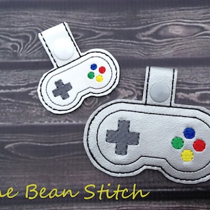 May include: Two grey and white embroidered game controller keychains with a button loop. The top keychain is white with grey details and the bottom keychain is silver with grey details. Both keychains have coloured buttons.