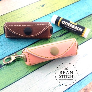 Könnte beinhalten: Zwei kleine Lederetuis mit Druckknöpfen und einer Metallschlaufe. Ein Etui ist braun und das andere rosa. Die Etuis sind für Lippenbalsam gedacht. Der Text "Otterbalm" ist auf einer Lippenbalsamtube sichtbar.