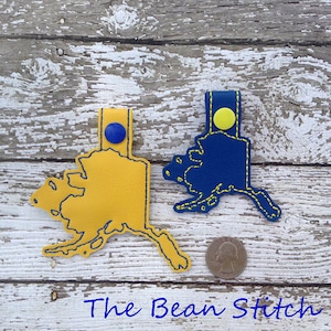 May include: Two leather keychains shaped like the state of Alaska. One is yellow with a blue button and the other is blue with a yellow button. Both have yellow stitching around the outline.