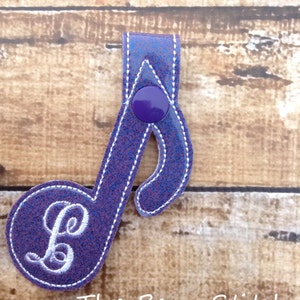 Può includere: Applicazione a forma di nota musicale viola glitterata con la lettera "L" ricamata in bianco. L'applicazione ha un bottone e cuciture bianche sui bordi. Il testo "The Bean Stitch" è in basso.