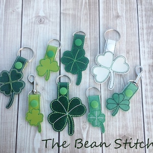 May include: A set of eight green and white shamrock keyrings with silver key rings. The keychains are made of felt and have a stitched design. The text "The Bean Stitch" is visible at the bottom of the image.