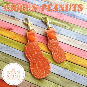 May include: Two orange leather peanut-shaped keychains with gold clasps and orange stitching. The keychains are on a multi-colored wooden background. The text "CIRCUS PEANUTS" is at the top of the image. The text "the BEAN STITCH embroidery design" is at the bottom of the image.