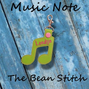 May include: Green music note keychain with the name "Londyn" embroidered in pink thread. The keychain has a silver metal clasp and a pink button. The keychain is on a blue wooden background with the text "The Bean Stitch" below it.