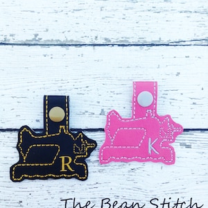 Könnte beinhalten: Zwei Leder-Nähmaschinen-Schlüsselanhänger, einer schwarz mit einem goldenen "R" und einer rosa mit einem weißen "K".