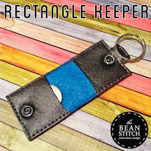 May include: A black leather rectangle keychain with a blue fabric pocket and a silver key ring. The keychain has a snap closure and the text "the BEAN STITCH embroidery design" is visible in the lower right corner.